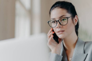 Hoş görünümlü esmer Avrupalı kadının yakın plan görüntüleri şeffaf gözlük takıyor, dizüstü bilgisayara odaklanmış, uzaktan çalışma sırasında telefonla konuşuyor, proje üzerinde çalışıyor. Teknoloji konsepti