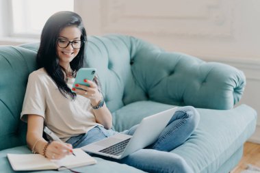 Yetenekli kız öğrenci proje için sunum yapıyor, cep telefonuyla gerekli bilgileri arıyor, dizüstü bilgisayarı açıyor, günlük giysiler giyiyor, rahat bir odada kanepede poz veriyor, internette sohbet ediyor.