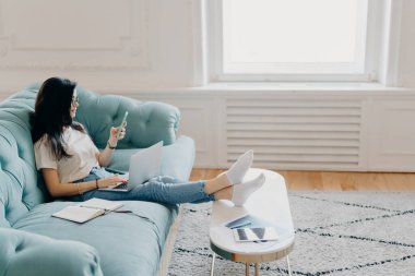 Rahat kız öğrencilerin yan fotoğrafları ev ödevi yaparken modern teknolojileri kullanır, proje yapmak için web kaynaklarını kullanır, rahat bir koltuğa oturur, masaüstüne yaslanır.