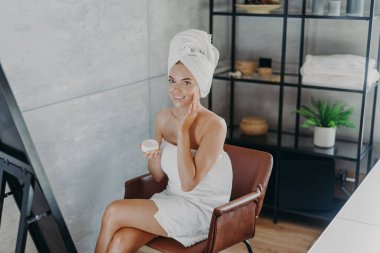 Gülümseyen rahat bir kadın güzel tenini önemser, rahat bir koltuğa oturur, rahat bir banyoda poz verir, aynada kendine bakar, cilt kremi kavanozunu tutar, nazikçe yanağına dokunur, havluya sarılır.