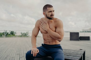 Düşünceli sakallı adam yoğun kardiyo eğitiminden sonra dinleniyor, eli göğsünde ve nefes darlığı çekiyor, dışarıda oturuyor, konsantre oluyor, üstsüz poz veriyor. Spor ve sağlıklı yaşam tarzı