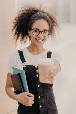 Kıvırcık saçlı güzel bir bayanın dikey pozu, hazır kahveyi elinde tutuyor, üniversitede molada içki içiyor, gözlük ve kot tulum giyiyor, olumlu duygular ifade ediyor. İçkili mutlu öğrenci.