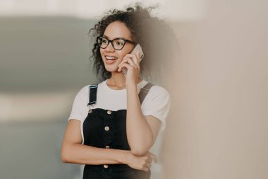 Hafif bir gülümseme ile gülümseyen genç kıvırcık kadın, cep telefonu görüşmeleri, dostça konuşma vardır, rahat beyaz tshirt ve tulum giyer, kenara odaklanmış. Boş zaman, yaşam tarzı, teknoloji kavramı