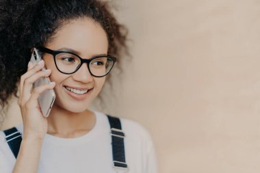 Cep telefonu ile afro saç görüşmeleri ile mutlu karanlık tenli kadın çekim yakın, nazikçe gülümsüyor, gözlük ve beyaz t shirt giyer, iyi haber alır, kahverengi arka planda izole, boş alan