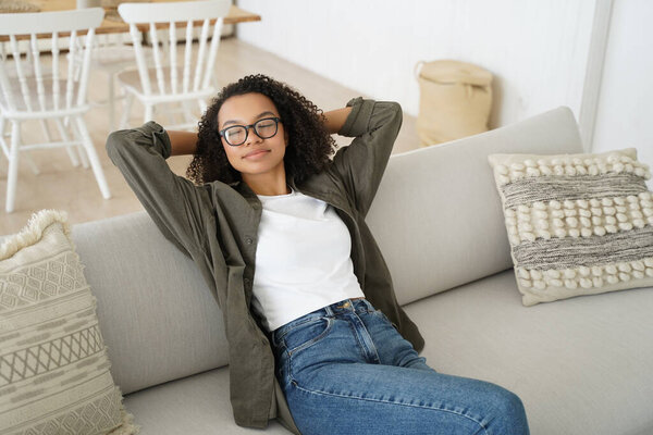 Спокойная молодая смешанная раса девушка опираясь на уютный диван с закрытыми глазами отдыхает дома, спокойная леди держит руки за голову, расслабляясь сидя на удобном диване в гостиной, наслаждаясь ленивыми выходными