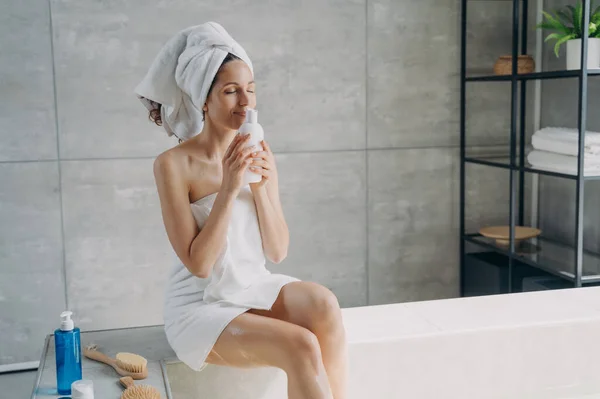 Relaxed happy woman tastes the fragrance of skincare spa cosmetic product sitting in bathroom. Female enjoying aroma of natural organic cosmetics for body care holding mockup bottle.