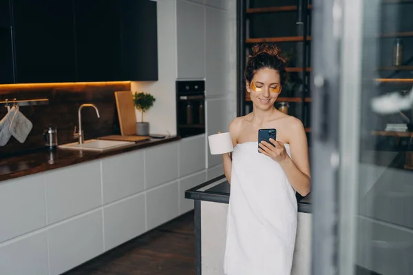 Beautiful woman with under eye patches, using smartphone on kitchen. Smiling girl wrapped in towel drinking coffee reading online news on social media during skincare routine after morning shower.