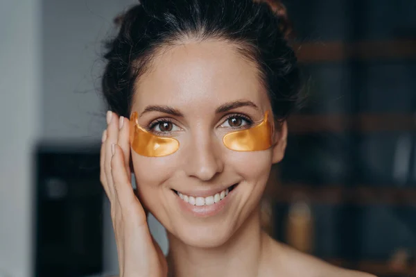 Smiling young latina woman applying moisturizing collagen golden under eye patches, happy pretty female does facial anti wrinkle beauty procedure at home, close up portrait. Skincare routine.