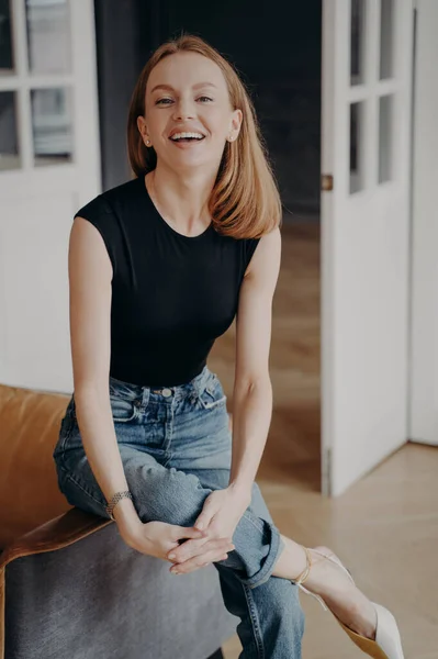 Lady is sitting on an armchair armrest and laughing. Emotional portrait of slim blonde tender woman. Redhead gorgeous lady in casual outfit is smiling. Happy european woman in apartment.