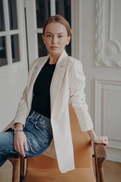 Contemporary gorgeous businesswoman is sitting on chair armrest. Portrait of lovely young adult european woman in white jacket posing in apartment. Concept of elegance, confidence and femininity.