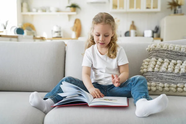Little caucasian girl is sitting on couch and reading fairy tale book. Kid is studying at home on quarantine. Preschool child is learning to read. Leisure and study at cozy home on quarantine.