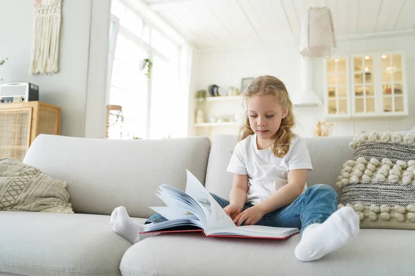 Lovely preschool child is learning to read. Little european girl is sitting on sofa and reading book. Kid is studying at home on quarantine. Leisure and study at cozy home on quarantine.