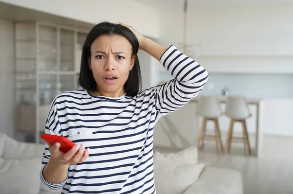Girl gets problem with phone. Worried woman gets unhappy news in message. Conceptual portrait of young emotional woman getting in trouble. Pretty hispanic lady in striped casual shirt at home.