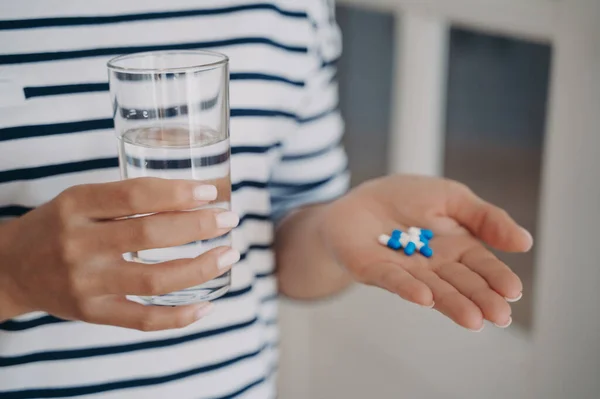 Hand of european woman is holding white and blue capsules and glass of water. Lady is going to take drugs. Therapy with medicine. Supplements and vitamins, pharmacology, influenza curation.