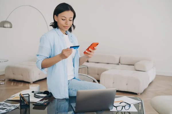 Happy spanish lady with credit card. Young woman is sitting on desk and typing password on smartphone. Online shopping, making order from home office on phone. Internet banking mobile app.