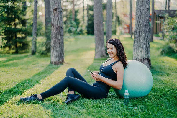 Relaxed sporty young female model takes break after aerobics exercises with fitness ball, sits on green grass, uses mobile phone, chats online, listens music in earphones, has athletic body.