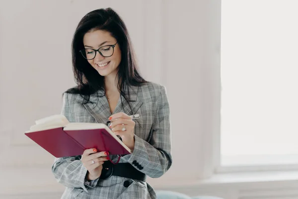 Horizontal shot of good looking woman professor writes down text notes in notepad, prepares for lecture in university, wears transparent glasses, business suit, stands indoor with broad smile