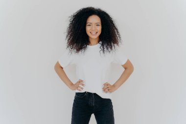 Keyifli kıvırcık kadının fotoğrafı iki elini de belinde tutuyor, nazikçe gülümsüyor, ince fiziği var, beyaz tişört giyiyor ve siyah kot pantolon giyiyor, keyfi yerinde, beyaz arka plana karşı kendinden emin duruyor.