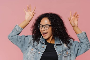 Kaygısız overjoyed koyu tenli genç kadın mutluluk dansları, eğlenceli kapalı, kollarını kaldırdı tutar, zevk gözleri kapatır, gözlük ve kot ceket giyer, pembe arka plan üzerinde izole.