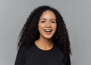 Mutlu koyu tenli Afro Amerikalı kadının yakın çekim olumlu gülüyor, iyi bir ruh hali içinde olmak, siyah rahat giysiler giymiş, gri arka plan üzerinde izole. İnsan duyguları ve hissetmek kavramı.