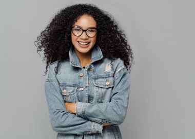 Gülümseyen kıvırcık bir kadın, nazik bir gülümseme, iyi bir ruh hali içinde, gözlük takıyor, kollarını kavuşturuyor, kot elbise giyiyor, kameraya düzgün bakıyor, güzel sohbetlerden hoşlanıyor, gri arka planda izole edilmiş.
