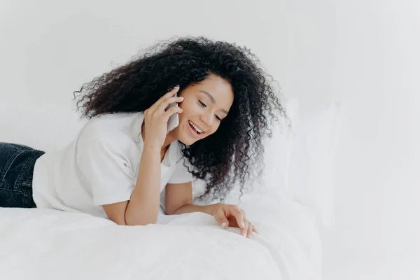 Pleased relaxed woman with curly hair has pleasant telephone conversation, speaks with friend during weekend before sleep, wears white t shirt, lies in cozy bed, keeps modern gadget near ear