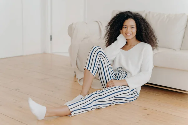 Ethnic girl feels relaxed and satisfied, sits on floor near comfortable sofa in empty room, wears white sweater, striped pants and socks, enjoys domestic atmosphere, enjoys coziness and comfort