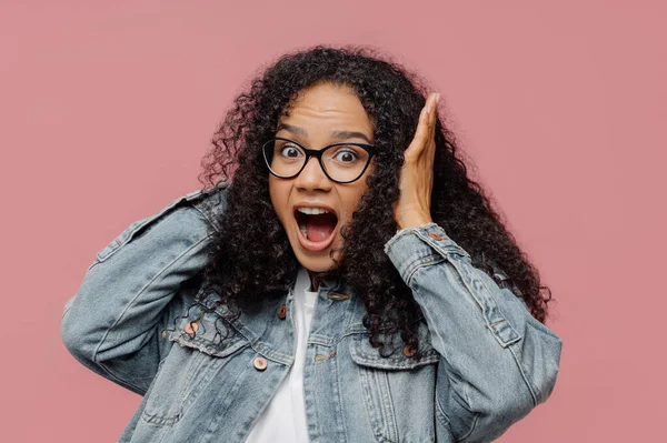 Photo of surprised Afro American woman covers ears, yells loudly, ignores loud sound, keeps mouth widely opened, wears spectacles and denim jacket, stands against rosy wall. Omg, how noisy there