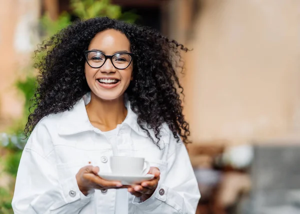 Outdoor shot of happy dark skinned lady with crisp hair, wears white jacket, drinks hot tea, has stroll across street, happy expression. Satisfied Afro woman enjoys spare time during weekend