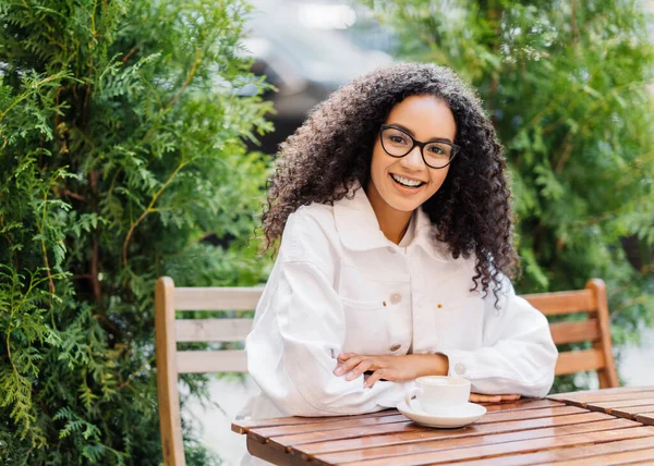 Relaxed cheerful Afro woman in white clothes, drinks coffee in outdoor cafeteria, sits at wooden table on chair against green thuja, has happy expression. People, lifestyle, spare time concept