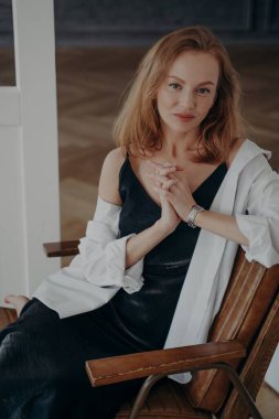 Siyah gece elbisesi ve beyaz gömlekli, kendine güvenen kızıl saçlı kadın. Gösterişli orta yaşlı bayan bir koltukta oturuyor. Mutlu zengin Avrupalı kadın bir partiye gidiyor. Moda kıyafetleri..