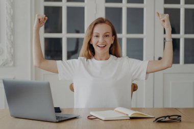 Kadın gücü işaret eder. Genç iş kadını dizüstü bilgisayarda çalışıyor. Beyaz tişörtlü mutlu Avrupalı kadın dairesinde uzaktan kumandayla çalışıyor. Sıkı çalışma ve başarı kavramı.