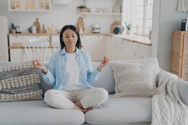 Sakin kadın yoga yapıyor, meditasyon yapıyor, nilüfer çiçekli koltukta oturuyor ve evde gözleri kapalı oturuyor. Huzurlu kadın zihinsel denge için nefes egzersizleri yapıyor. Sağlıklı yaşam tarzı, endişe giderici.