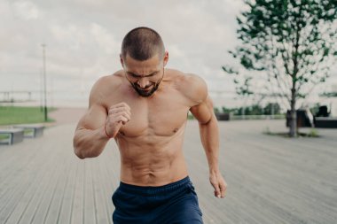 Sportif sakallı erkek koşucu nehir ya da denizin yanındaki yürüyüş yolunda koşar, günlük antrenman yapar, çıplak gövdeyle poz verir, tüm çabalarını sağlıklı ve formda kalmaya harcar. Sabah sporu konsepti