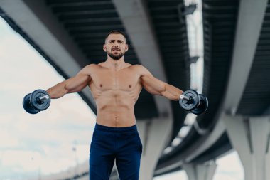 Kaslı vücutlu güçlü sakallı genç adam ağır halterlerle kollarını kaldırıyor spor kıyafetleri giymiş spor malzemelerini açık havada kullanıyor. Sağlıklı yaşam tarzı egzersizi ve vücut geliştirme konsepti