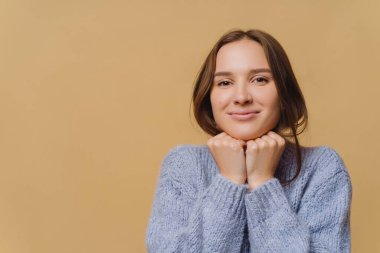 Sağlıklı ciltli, iki elini de çenesinin altında tutan, büyük boy kazak giyen, doğrudan kameraya bakan, kahverengi stüdyo duvarının üzerinden izole edilmiş hoş, koyu renk saçlı bir kadın. İnsanlar, insan yüz ifadeleri..