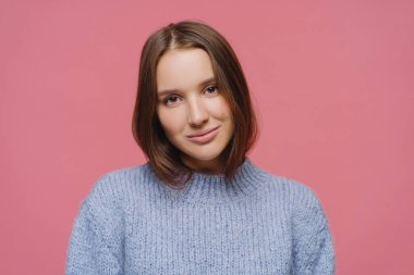 Tatmin olmuş koyu renk saçlı genç bayanın yakın çekimi başını eğiyor, hoş sohbetlerden hoşlanıyor, sıcak kış süveteri giyiyor, pembe arka planda tek başına poz veriyor, kameraya odaklanmış, rahatlamış hissediyor.