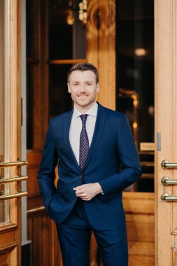 Resmi takım elbise giymiş, sakallı yakışıklı bir erkeğin dikey portresi, finans projesiyle uğraşırken tatmin olmuş, ofisin girişinde duruyor. İş konsepti