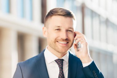 Kişi, iş ve kariyer kavramı. Yakışıklı neşeli başarılı erkek resmi kıyafetler, kıl ve olumlu gülümseme, işadamı, arka plan bulanık karşı pozlar ile akıllı telefon yolu ile konuşuyor