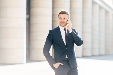 Neşeli erkek girişimci telefon görüşmesi yapar, kendine güveni tam ve mutlu bir şekilde bir kenara bakar, resmi kıyafetler giyer, operatör ya da iş ortağıyla konuşur, resmi takım elbisesinin cebini doldurur.