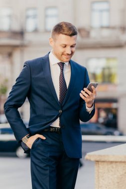 Zengin erkek yöneticinin dikey görüntüsü şık bir takım elbise giyer, elini cebine sokar, akıllı telefon kullanır, sürekli iletişim halinde olur, internet sitesinde iş haberleri okur, ekrana konsantre olur..