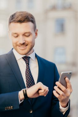 Keyifli tıraşsız erkek şirket sahibi kol saatine mutlu bir şekilde bakar, saati kontrol eder, akıllı telefondan gelen mesajları alır, banka hesabının profilini günceller, ücretsiz bağlantı kullanır