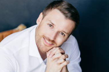 Çekici genç tıraşsız Kafkas erkek ile anız, portre pozitif bir görünüme sahiptir, çene, koyu arka plan üzerinde izole zarif beyaz elbise giymiş ellerimde tutar. Başarılı bir iş adamı