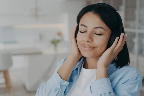Calm young female wearing wireless headphones listening to relaxing music, enjoying sound at home. Happy woman listens to audio affirmations or meditation, resting with closed eyes at home.