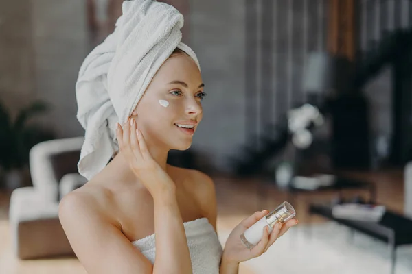 Beautiful young European woman with white bath towel on head, applies face cream and body lotion, enjoys skin care procedures after shower, poses at home alone. Daily beauty routines concept