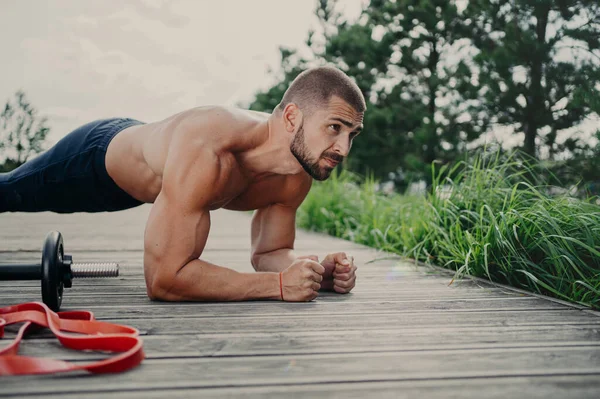 Muscular bodybuilder stands in plank pose demonstrates endurance has strong biceps poses outdoor has athletic workout, poses outdoor, keeps good physical shape. Man doing push ups, concentared forward