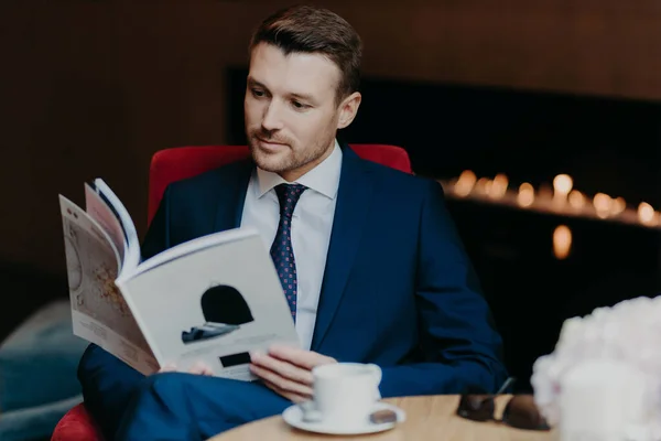 Serious male entrepreneur reads business magazine, sits in coffee shop, drinks coffee or tea, dressed in formal suit, has dinner break and rest after being in office. Business and lifestyle concept