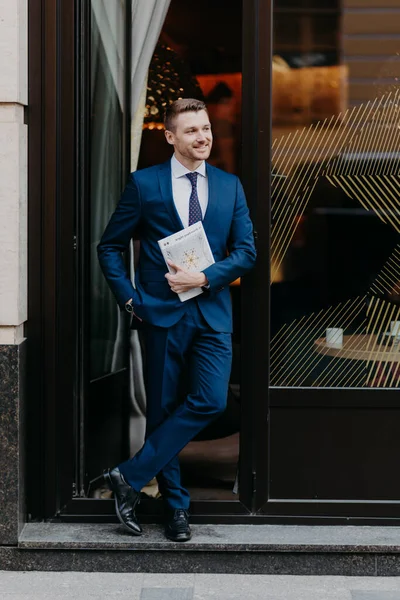 Confident successful businessman in formal suit, keeps hand in pocket, holds business magazine, stands near entrance of cozy cafeteria, has pleased expression. People, job and occupation concept
