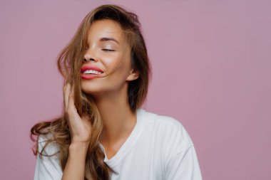 Uzun sarı saçlı rahat güzel bir kadın, gözlerini zevkten uzak tutuyor, olumlu gülümsüyor, fotoğraflanmaktan hoşlanıyor, kameraya poz veriyor, makyajının mor stüdyo duvarında olduğunu gösteriyor.