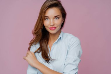 Hoş görünümlü uzun saçlı bir kadın modelin portresi, kameraya özgüvenli bir şekilde bakıyor, makyaj yapıyor, nazikçe gülümsüyor, günlük kapağı için poz veriyor, mor arka planda izole edilmiş..